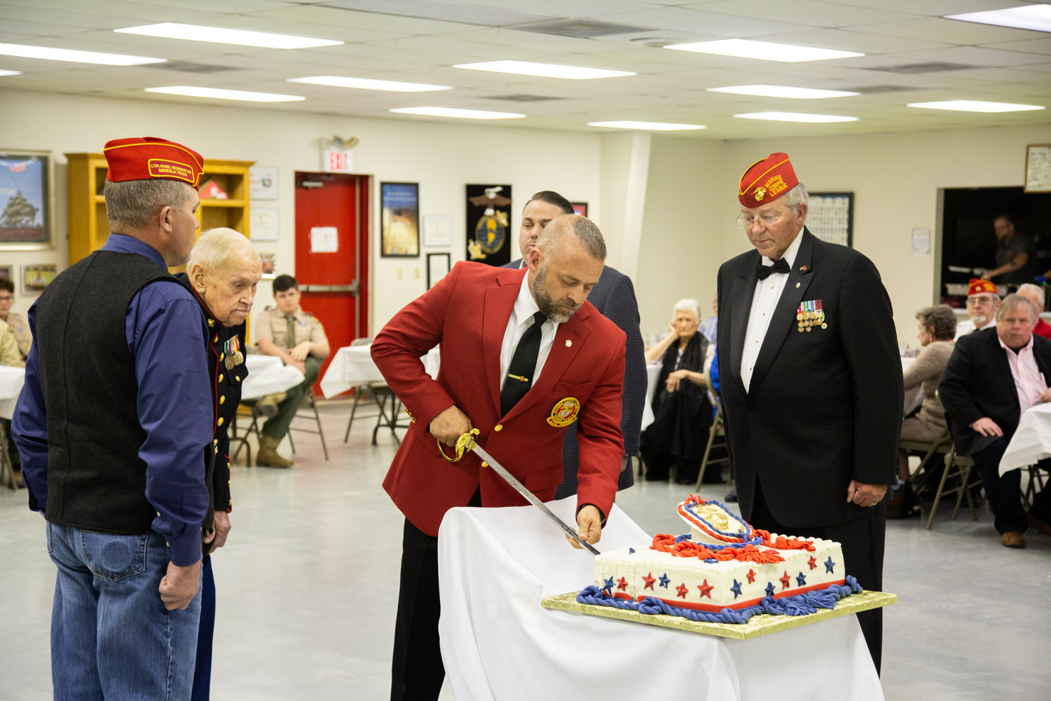 Marine Corps League Commandant Kelly Smith cuts the Marine Corps birth- day cake with the traditional Mameluke sword. The oldest Marine present, PFC James Krodel, and youngest Marine present, Lance Corporal Brandon Baade, watch with cake escorts James Biesheuvel and Mike Jernigan.